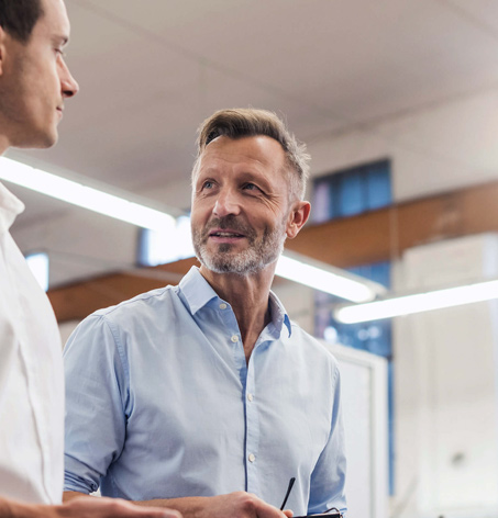 Two men are engaged in conversation indoors, with one holding a pair of glasses. Both are wearing light-colored shirts and appear to be in a well-lit office or industrial space.