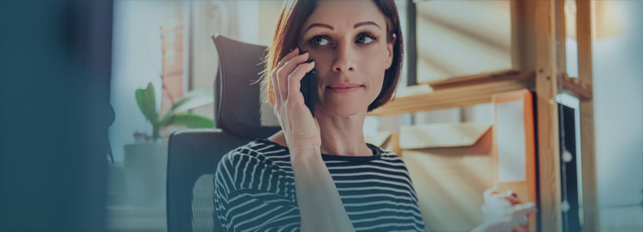 A woman with a short bob haircut is seated and talking on a phone. She is indoors, wearing a black and white striped shirt, with blurred office or home office elements in the background.