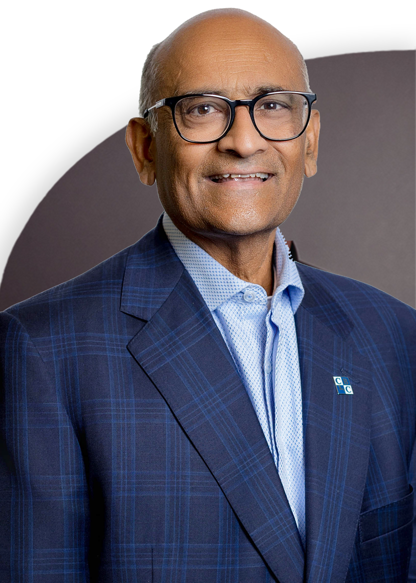 An older man in a checkered suit and blue shirt smiles at the camera. He is wearing glasses and has a badge on his lapel.