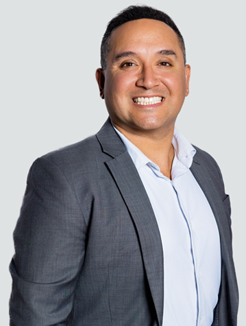 A person in a gray suit and light blue shirt smiles at the camera against a plain background.