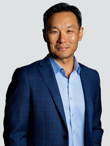 Man in a blue suit and light blue shirt smiles at the camera against a plain background.