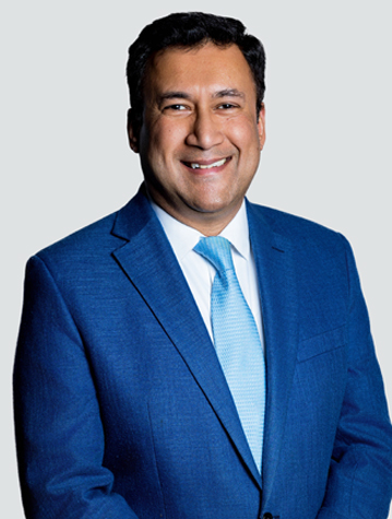 A person in a blue suit and light blue tie smiles against a plain background.