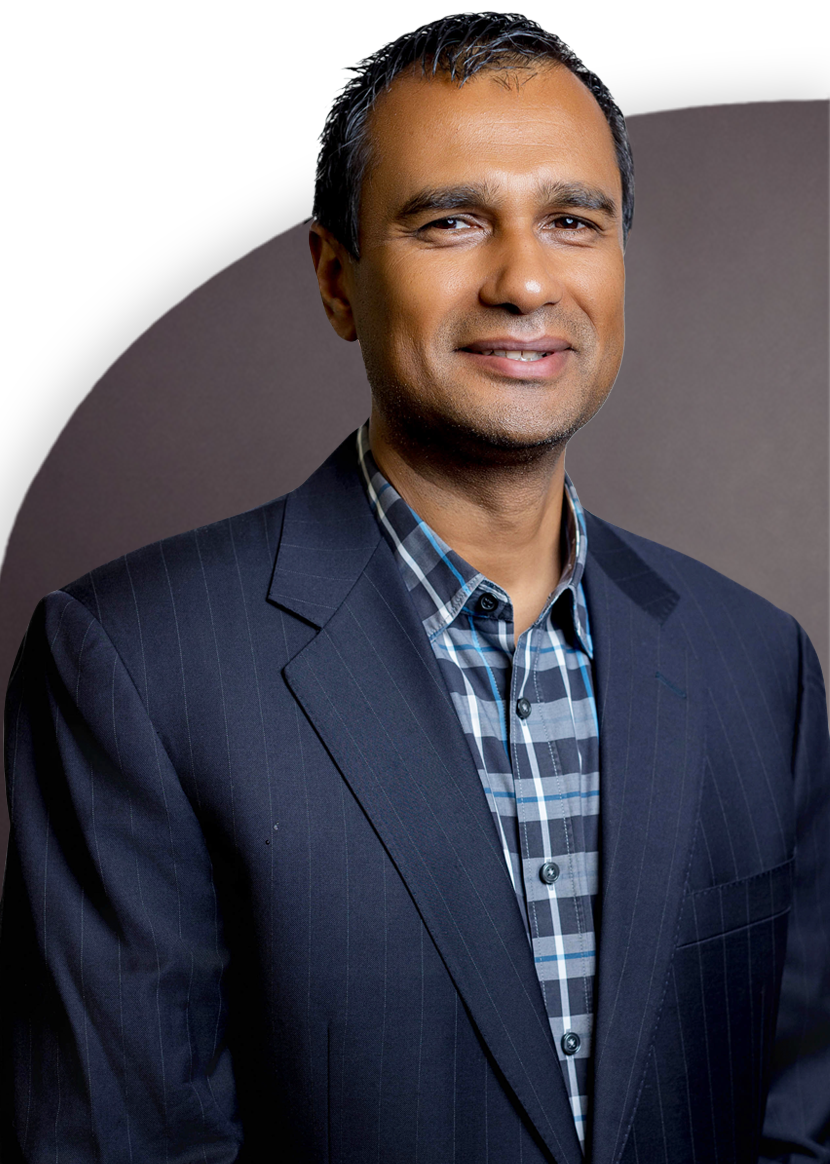 A person wearing a dark suit and a plaid shirt, smiling, with a neutral background.