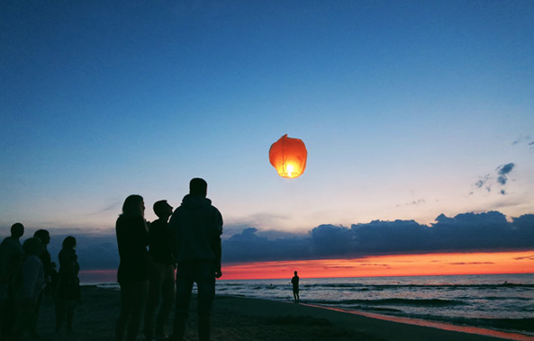 A group of people on a beach watches a single illuminated lantern floating in the sky during sunset.