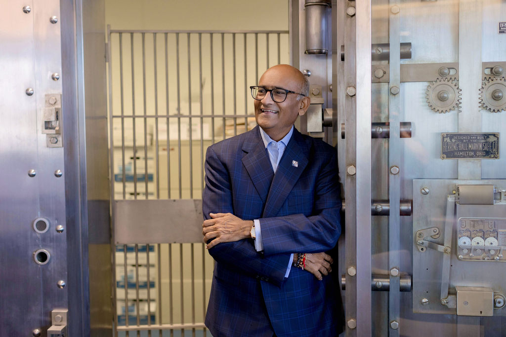 A man in a suit stands smiling with arms crossed inside a bank vault, with visible gears and a barred door in the background.