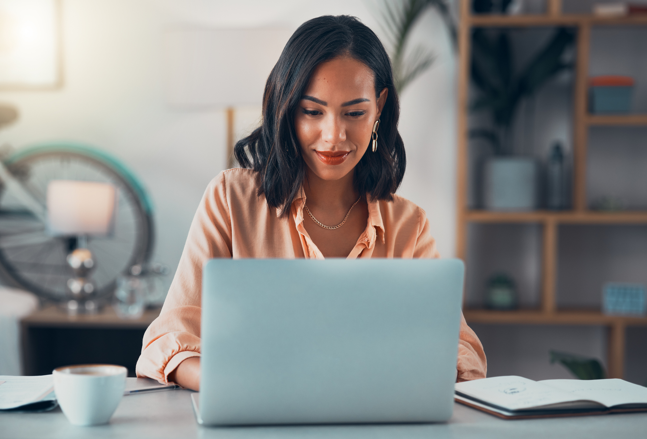 Woman with shoulder-length hair working on a laptop at a desk, surrounded by books and a cup of coffee, with a bike and shelf in the background.