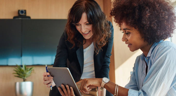 Two women are collaborating in an office, with one seated and pointing at a tablet that the other is holding.