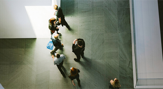 Aerial view of people walking in a modern building with large grey floor tiles and white walls, with one person near an illuminated area.