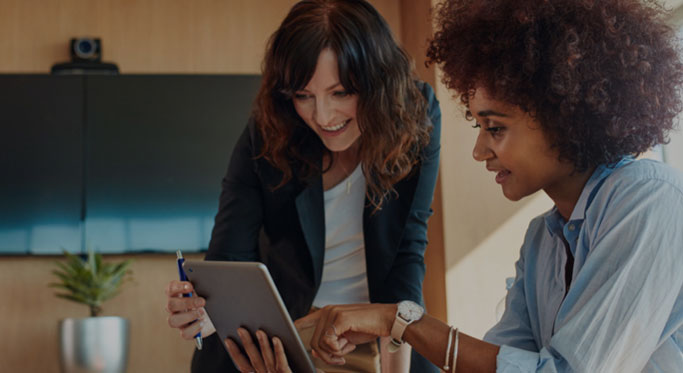 Two women looking at a tablet, one pointing at the screen while the other holds the device and smiles. They appear to be in an office setting with a TV screen and a plant in the background.