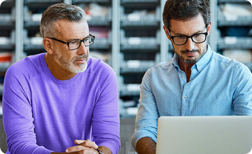 Two men, both wearing glasses, are seated and looking at a laptop. One man wears a purple shirt, and the other wears a light blue polka-dot shirt. Shelves with boxes are visible in the background.