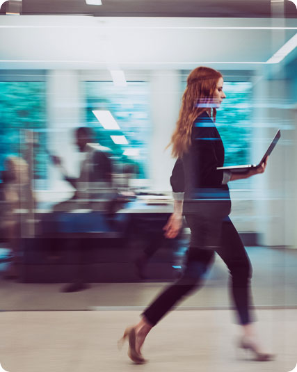 A woman in business attire walks briskly through an office while holding a laptop. Blurred background shows other office workers and large windows.