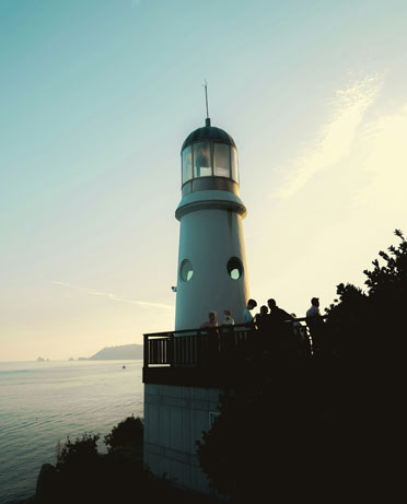 A group of people stand on a viewing platform next to a white lighthouse overlooking the ocean with a clear sky in the background.