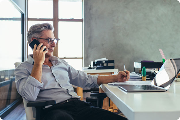 A man with gray hair and glasses sits at a desk in an office, talking on the phone while writing and looking at a laptop.