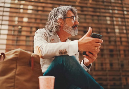 A man with gray hair and a beard sits on the ground, holding a phone and looking to the side. A brown bag and a disposable coffee cup are placed nearby.