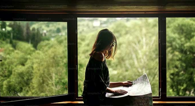 A person with long hair reads a map while sitting by a large window with a scenic view of green trees outside.