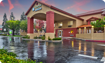 A hotel with a red brick facade and covered entrance sits in front of a wet parking lot and sidewalk after rain, under a sky with pink and blue hues. Trees and shrubs surround the building.