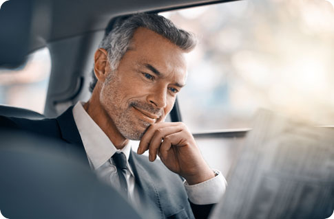 A man with gray hair and a beard, dressed in a suit, reads a newspaper while sitting in the backseat of a car, with sunlight streaming through the window.