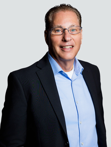 A man in glasses, wearing a black blazer and blue shirt, smiles at the camera against a neutral background.