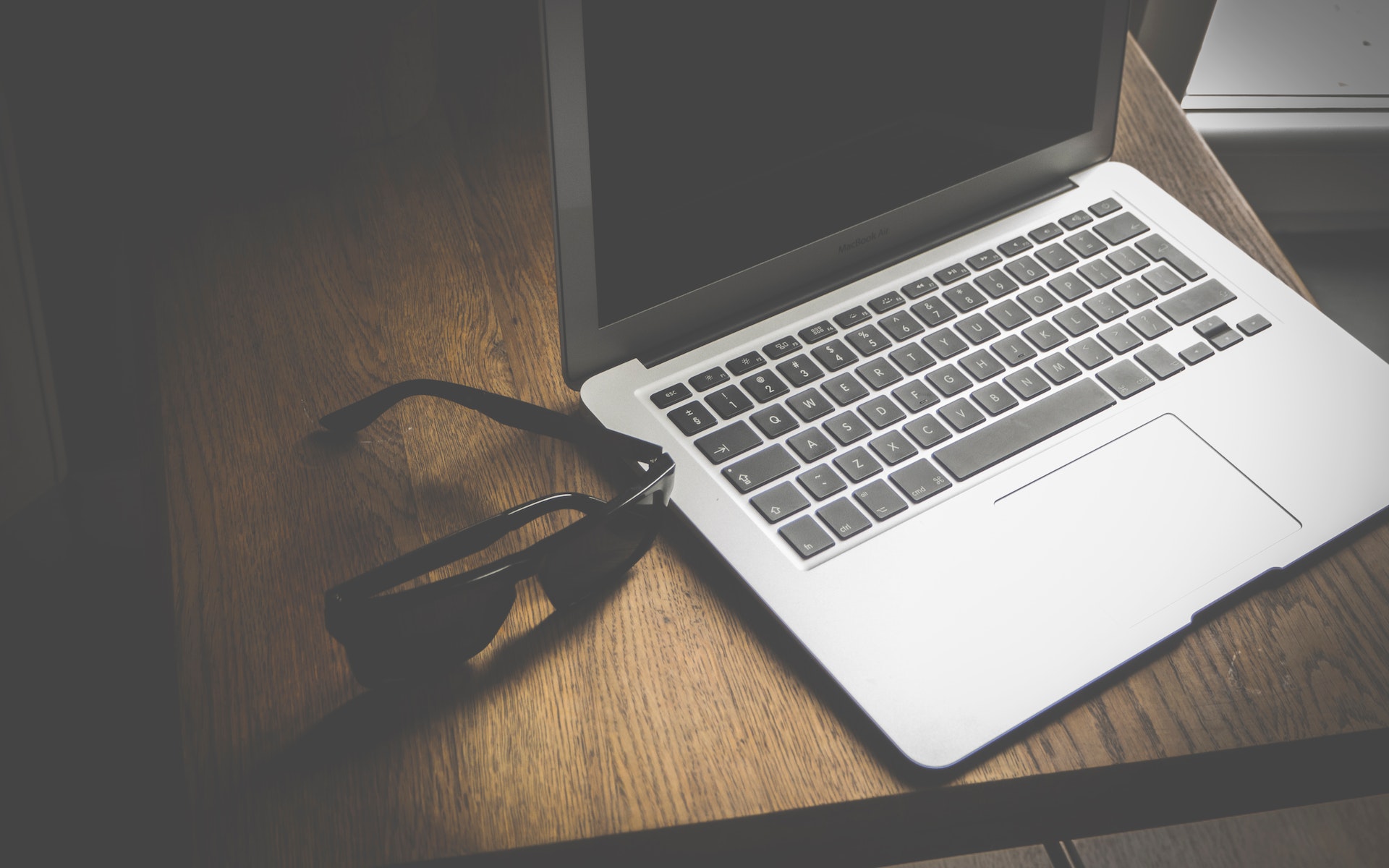 A silver laptop with a black screen sits open on a wooden desk next to a pair of black sunglasses.