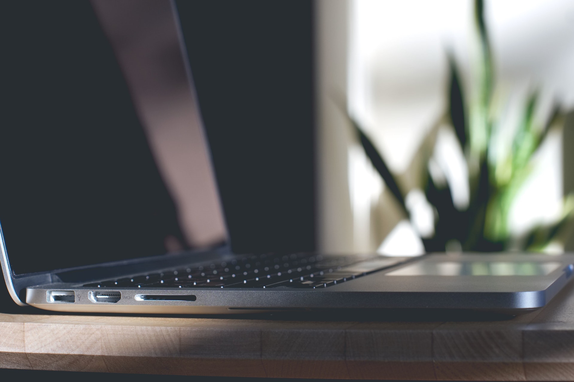 Close-up of a laptop on a wooden desk, showing the side ports and part of the keyboard. In the blurred background, there is a houseplant.