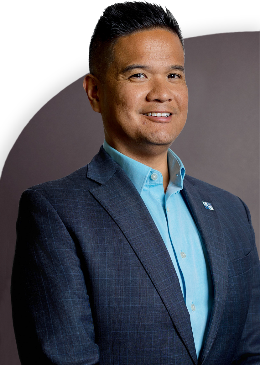 Man in blue suit and light blue shirt, smiling, stands against a dark background.