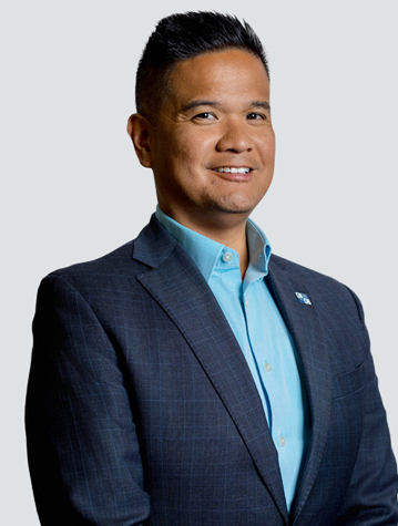 A person in a suit jacket and light blue shirt stands against a plain background, looking slightly to the side with a neutral expression.