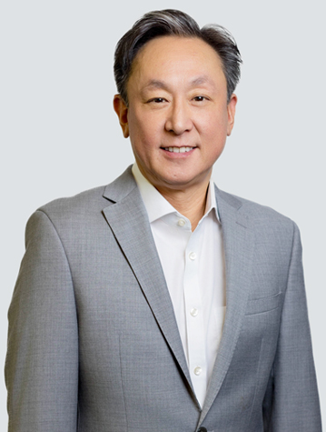 A man in a gray suit and white shirt stands against a light background, facing the camera.