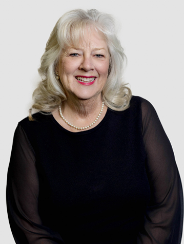 Elderly woman with long white hair, wearing a black top and pearl necklace, smiling against a plain background.