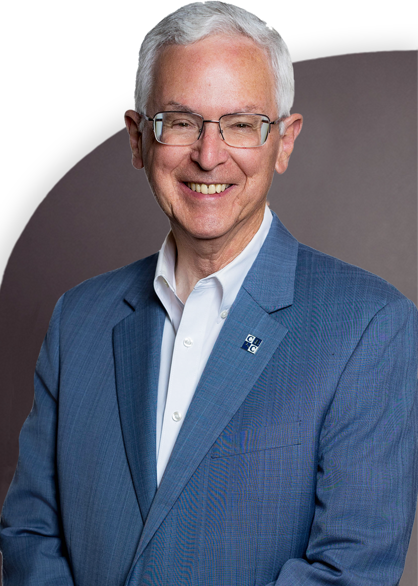 A smiling man with white hair and glasses in a blue suit and white shirt stands against a gray background.