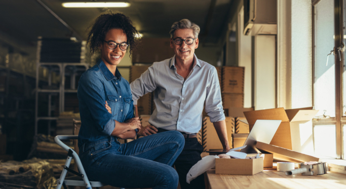 Two people in casual attire smiling and sitting in a workshop or warehouse space, with boxes and packing materials around them.