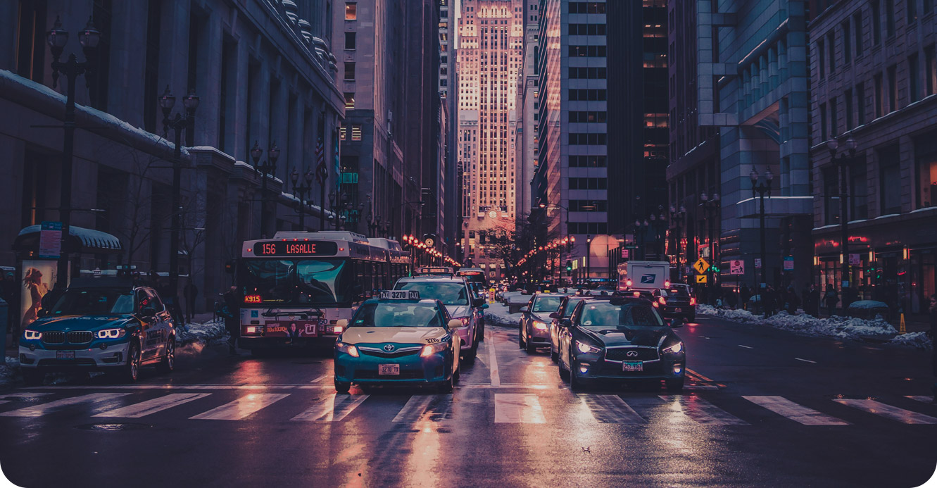 A busy city street at dusk with cars and a bus stopped at a crosswalk, surrounded by tall buildings with lights on.