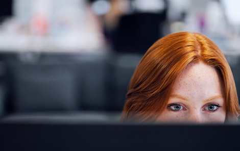 A person with red hair and freckles closely focused on a computer screen in an office setting.