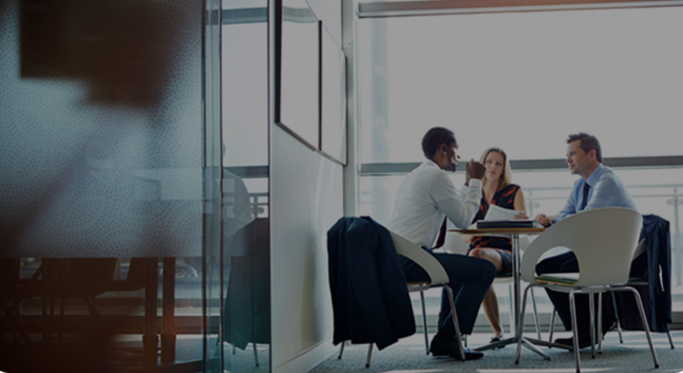 Three people sit at a table in a modern office environment, engaged in a discussion. A window in the background allows natural light into the room.