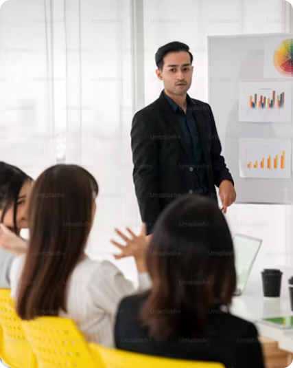 A man in a suit stands near a whiteboard with charts, addressing a group of people seated around a table.