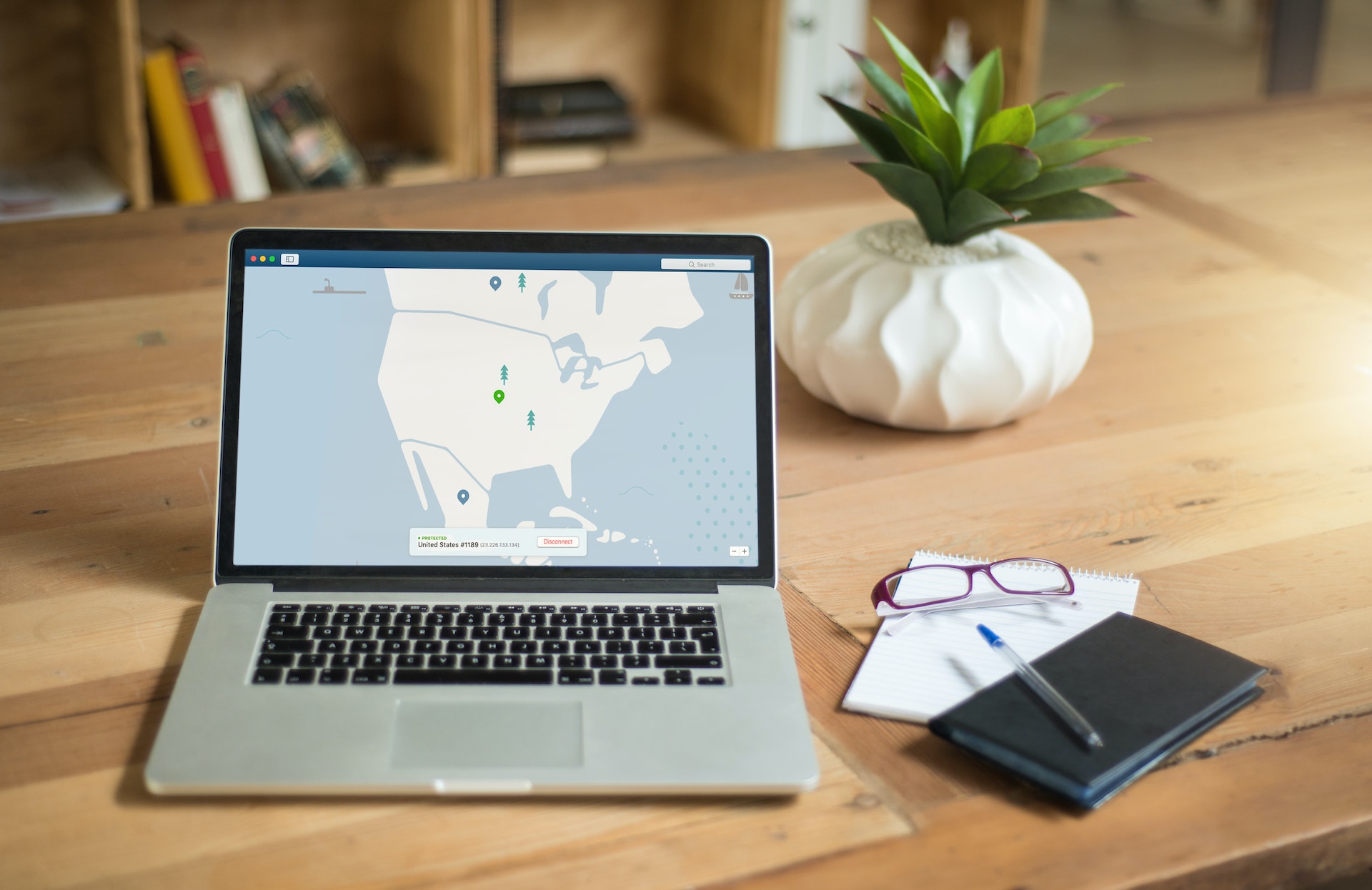 A laptop displaying a map is on a wooden table with a potted plant, eyeglasses, a notebook, and a pen beside it.