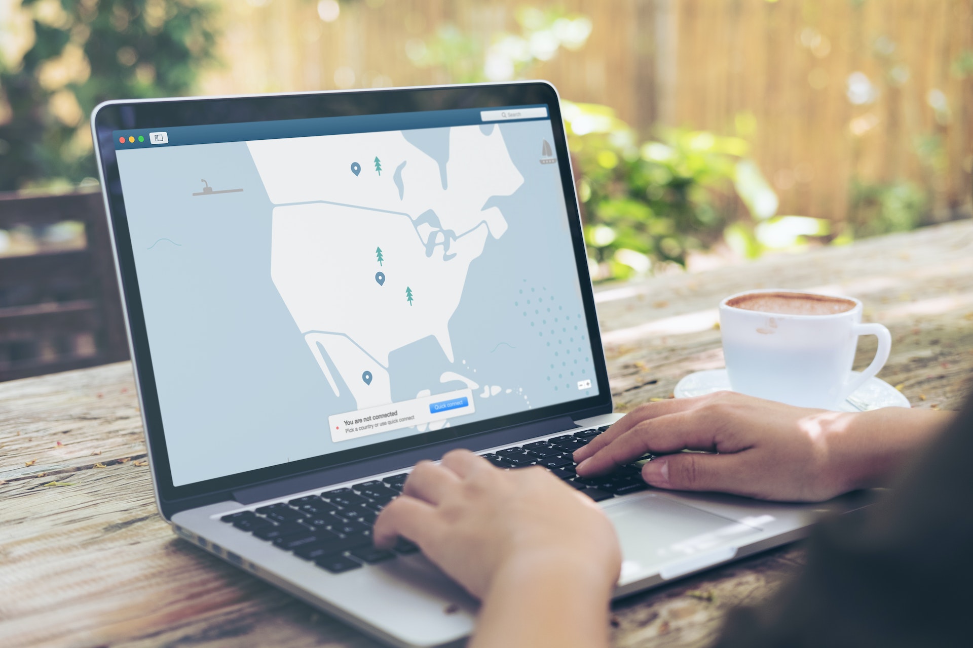 A person uses a laptop at an outdoor table with a cup of coffee nearby. The screen shows a map with several marked locations.