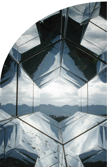 View of a cloudy sky and forested mountains through a geometric, mirrored structure, creating multiple reflections.