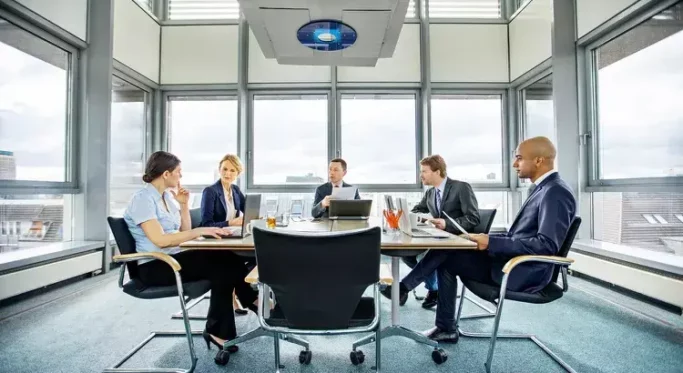 Five people in professional attire sit around a conference table in a modern office, engaged in discussion. Laptops and documents are on the table. Large windows offer a view of the cityscape outside.