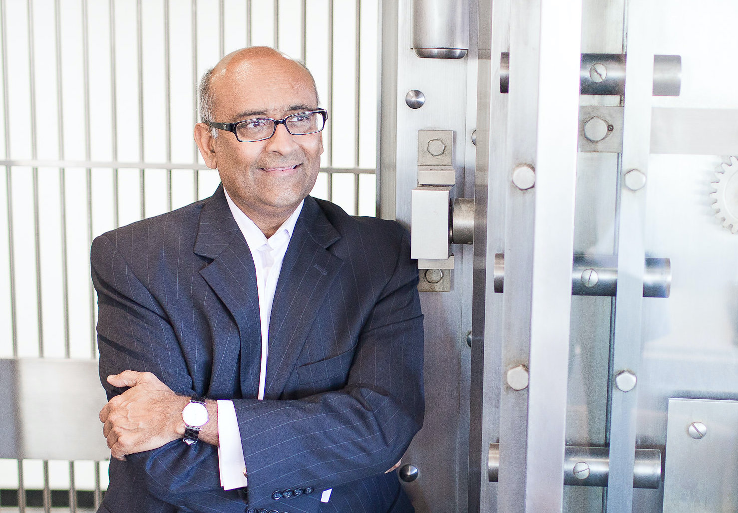 A man in a suit and glasses stands with his arms crossed, leaning against a large metal vault door.