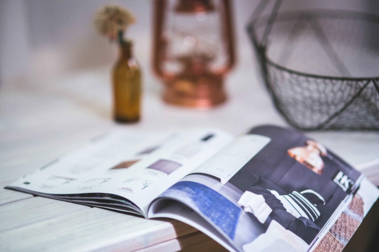 A magazine lies open on a wooden table next to a small amber bottle, a wire basket, and a copper lantern.