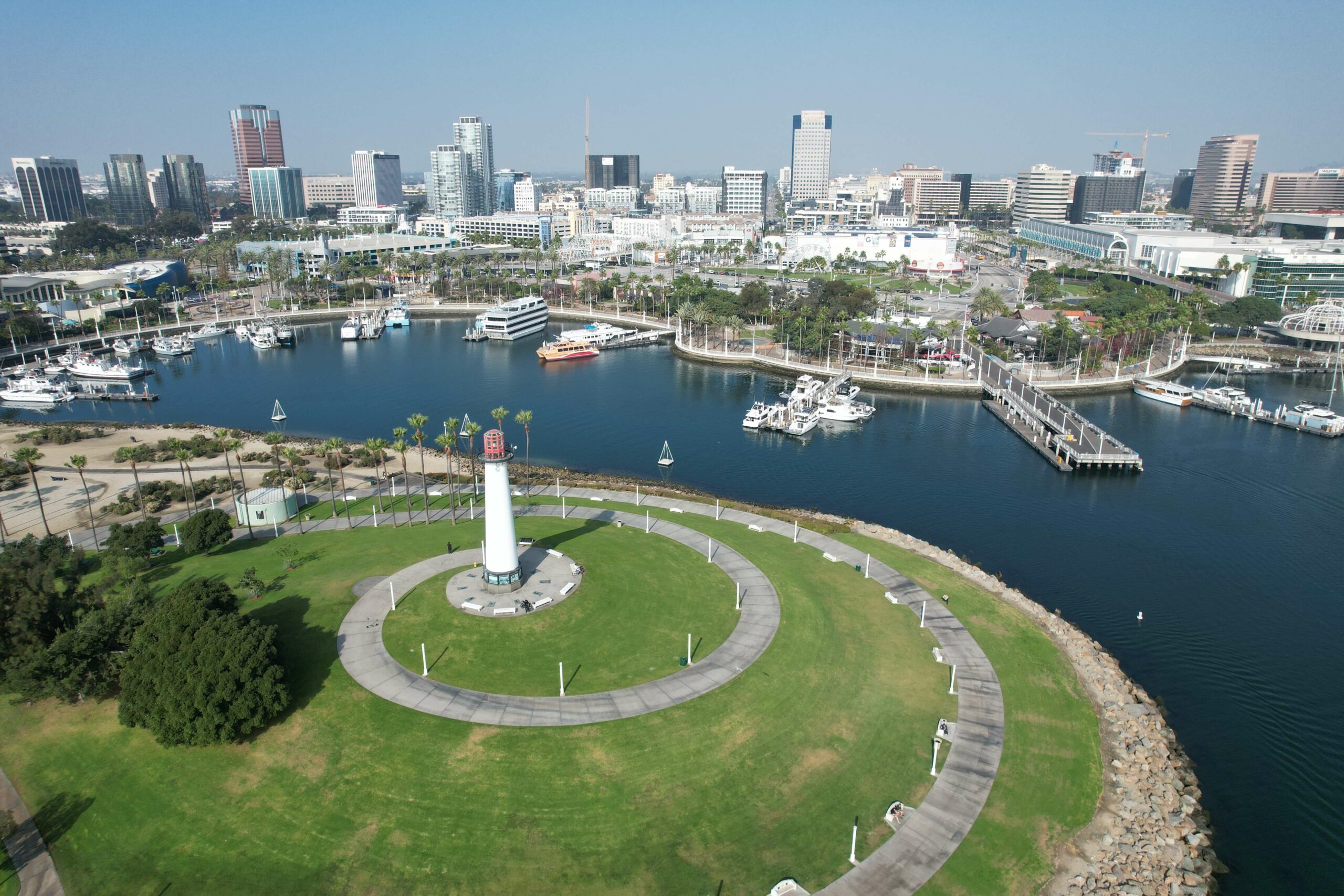 Aerial view of a coastal city with a central white lighthouse, surrounded by green grass, circular walkways, docks, boats, and numerous buildings in the background.