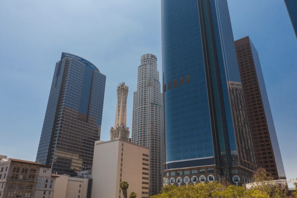 A cluster of modern skyscrapers with reflective glass windows and varied architectural designs against a clear blue sky. Trees are visible in the lower part of the image.