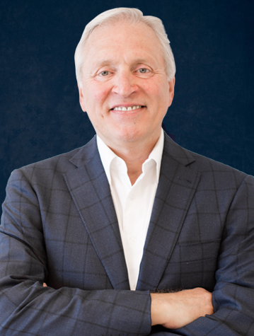 A man with white hair, wearing a dark, checkered suit jacket and white shirt, stands with arms crossed and smiles against a dark blue background.