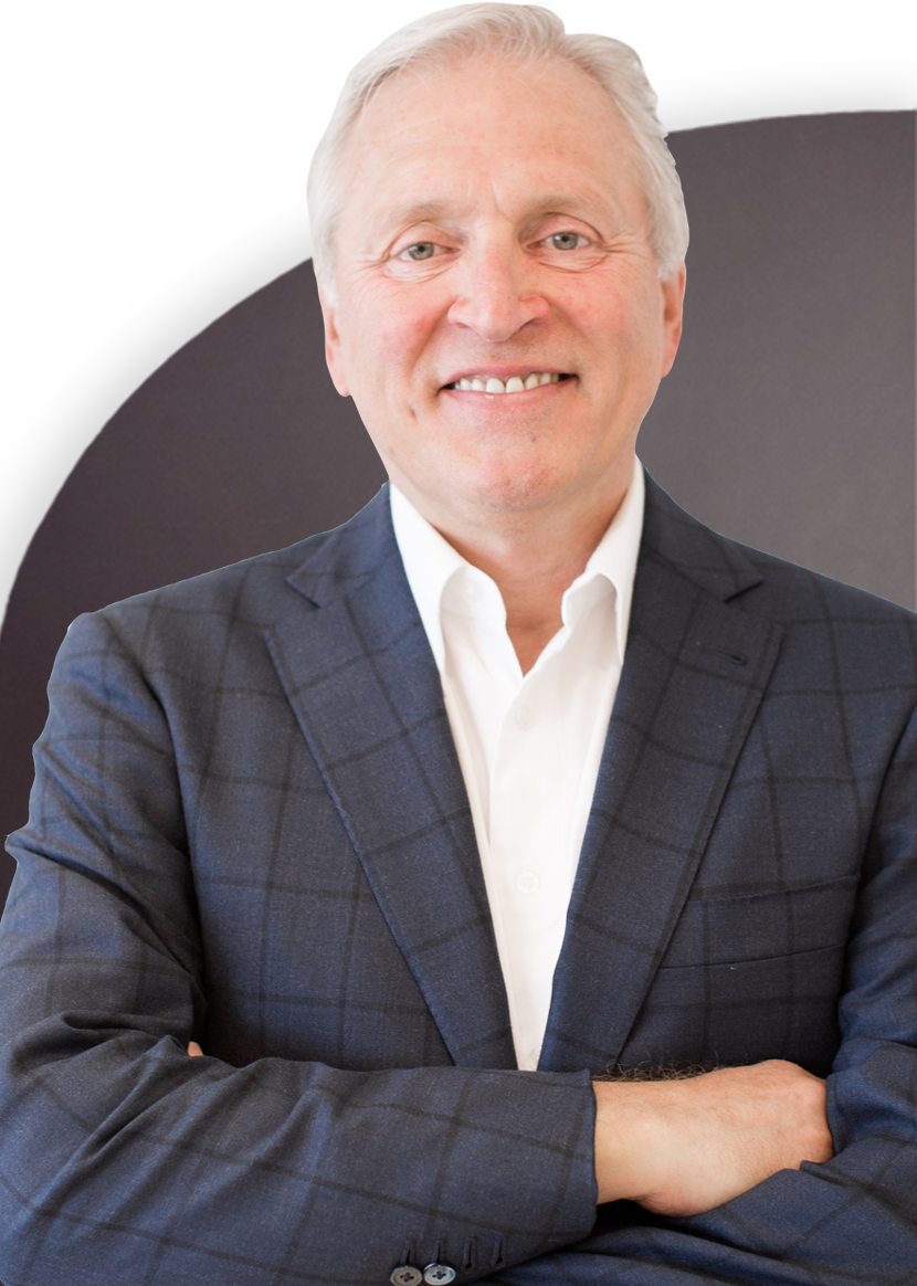 A man with grey hair wearing a dark suit and white dress shirt stands with arms crossed, smiling at the camera.