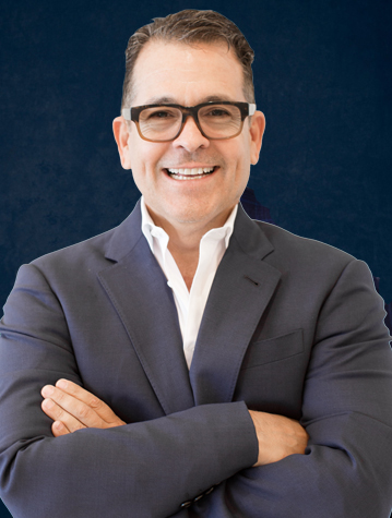 A man in a dark suit and white shirt smiles with his arms crossed in front of a dark blue background. He is wearing glasses.