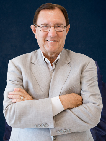 A man wearing glasses and a light gray suit stands with his arms crossed, smiling against a dark blue background.