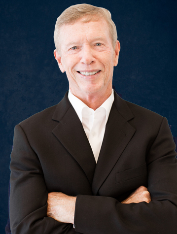 A smiling man with gray hair, wearing a black suit and white shirt, stands with his arms crossed against a dark blue background.