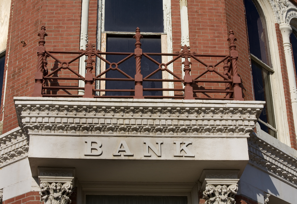 A red brick building with a white stone facade and the word "BANK" inscribed above the entrance. A decorative iron balcony and tall arched windows are visible.