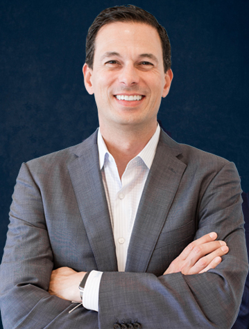 Man in a gray suit jacket and white shirt stands confidently with arms crossed against a dark blue background, smiling at the camera.