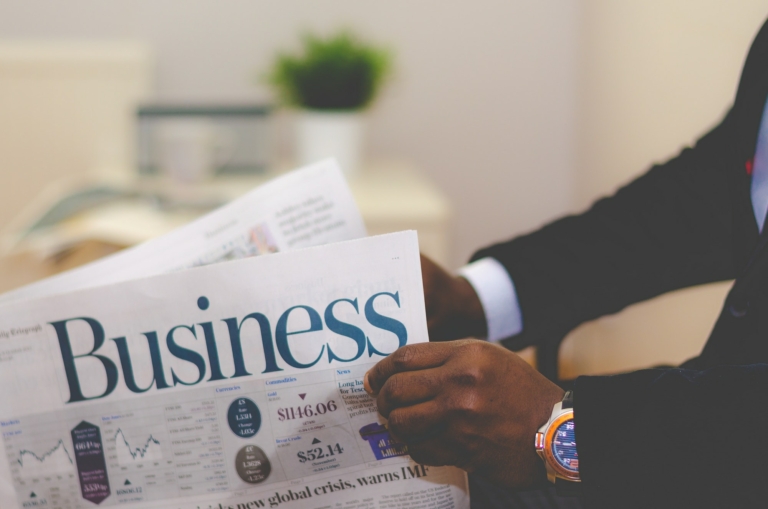 A person in a suit reads a newspaper with a "Business" section headline. A green plant and an open book are in the background.